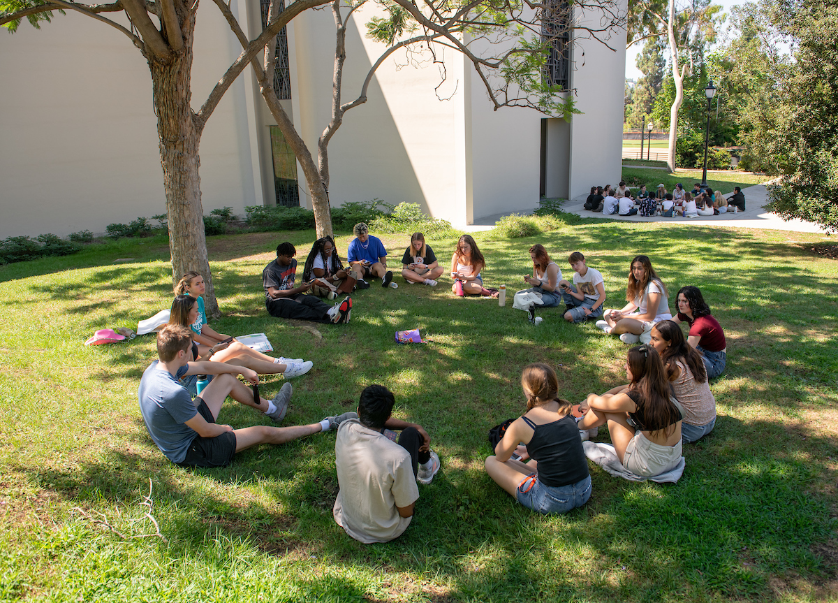 Student sit outside in a circle, under the shade of trees